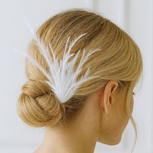 May include: A white feather hair clip is placed in the back of a person's blonde hair, styled in a low bun. The hair accessory is made of delicate, feathery plumes, creating a soft, elegant look. The person is wearing a white dress.