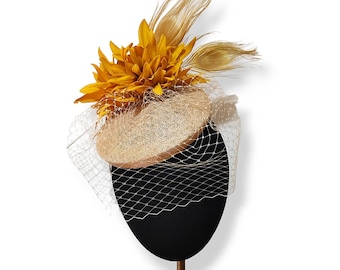 Puede incluir: Sombrero decorativo con base tejida color beige, adornado con una gran flor amarilla y plumas doradas. Una redecilla blanca cubre la parte delantera. El sombrero se exhibe sobre una forma ovalada negra con un soporte dorado.
