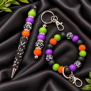 May include: A beaded pen, a beaded keychain, and a beaded wristlet featuring a mix of matte black, lime green, purple, and orange silicone beads, along with black beads patterned with white skulls and crossbones and silver rhinestone spacers.