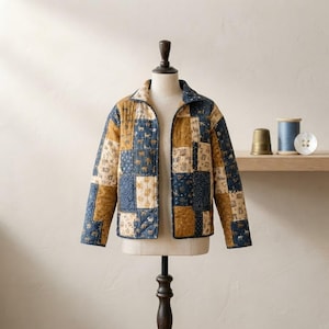 Puede incluir: Una chaqueta acolchada de manga larga con un diseño de patchwork de paneles rectangulares de tela en tonos azul marino, amarillo mostaza y crema. La tela está decorada con pequeñas ilustraciones repetitivas de animales y motivos botánicos.
