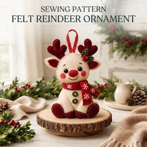May include: A felt reindeer ornament featuring the text SEWING PATTERN FELT REINDEER ORNAMENT. The cream-colored reindeer has dark red antlers, a red nose, and a red scarf with white snowflake patterns. It includes two green buttons on its chest.