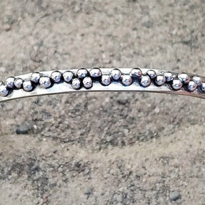 May include: A silver cuff bracelet with a beaded design. The bracelet features a row of small, round beads along the top edge. The open cuff design allows for easy wear. The bracelet is shown on a textured, light-colored surface.