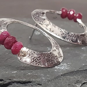 May include: Silver earrings with a textured oval design and three red gemstone beads. The earrings have a post back.