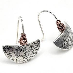May include: A pair of silver earrings with a half-moon shape and a copper wire accent. The earrings have a textured surface with a swirling pattern.