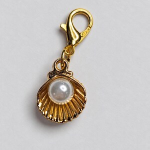 May include: A gold-toned metal charm featuring a scalloped seashell design with a central white spherical pearl. The charm is attached to a gold-toned lobster claw clasp, allowing it to be easily clipped onto various items or jewelry pieces.