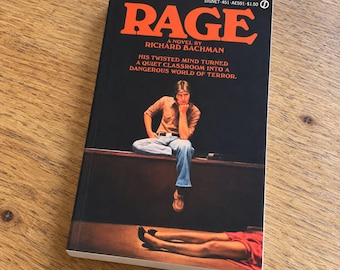 May include: A paperback book titled Rage by Richard Bachman with a black cover. The cover features large orange text, a person sitting on a desk, and a pair of legs in a red skirt lying on the floor. The text reads: His twisted mind turned a quiet classroom into a dangerous world of terror.