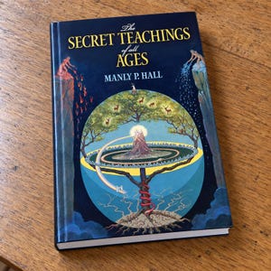 May include: A hardcover book titled The Secret Teachings of All Ages by Manly P. Hall. The dark blue cover features a central circular illustration of a tree with deer, a mountain, and a red snake coiled around the trunk, flanked by cliffs.
