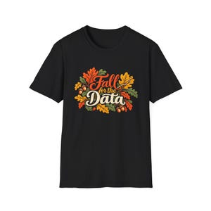Może przedstawiać: Czarny t-shirt z krótkim rękawem z centralną grafiką przedstawiającą kolorowe jesienne liście i żołędzie otaczające napis "Fall for the Data" stylizowaną czcionką, z dołączoną małą ikoną wykresu liniowego z trendem wzrostowym.