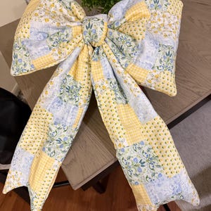May include: A large, quilted fabric bow featuring a patchwork design of yellow and white gingham, light blue floral patterns, and sprigs of lavender. The bow has two wide, long tails and a central knot, all constructed from soft, padded textile.