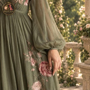 Puede incluir: Un vestido largo de color verde salvia con mangas largas y transparentes tipo obispo y cintura fruncida. La tela est&aacute; adornada con patrones florales bordados en rosa y rosa oscuro. Un logotipo circular en la esquina superior izquierda dice Morrow Wear.