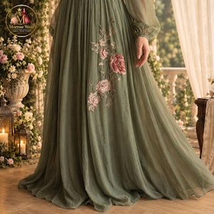 Puede incluir: Un vestido largo de color verde salvia con mangas largas transparentes y falda plisada. La tela est&aacute; adornada con un patr&oacute;n floral bordado en tonos rosa y crema, que cae por el lateral de la falda desde la l&iacute;nea de la cintura.