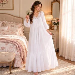 Puede incluir: Un camisón blanco largo hasta el suelo con mangas largas y holgadas, escote en V con delicados adornos de encaje y falda fruncida. La prenda está confeccionada en un tejido ligero y presenta una sutil textura plisada vertical en el corpiño.