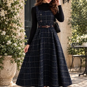 Puede incluir: Un vestido tipo pichi azul marino a cuadros con escote cuadrado, dos botones decorativos en el corpiño y un cinturón de cuero marrón en la cintura. El vestido se lleva sobre una camiseta negra de cuello alto y manga larga con botines negros.