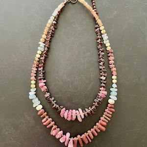 May include: A layered necklace with a mix of pink, red, and blue gemstone beads. The necklace is made of natural stones and has a rustic, bohemian style.
