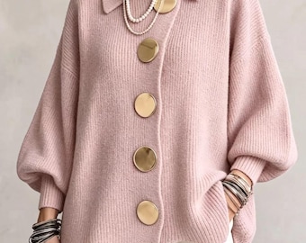 Könnte beinhalten: Eine hellrosa Strickjacke aus Rippstrick mit Kragen, langen Ärmeln mit gerippten Bündchen und vier großen, runden, goldfarbenen Metallknöpfen auf der Vorderseite. Das Kleidungsstück wird mit einer weißen Hose und Perlenketten kombiniert.