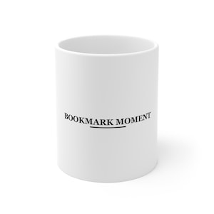 Peut inclure: Un mug en céramique blanche avec le texte noir BOOKMARK MOMENT imprimé en police serif sur le devant, accompagné d'une ligne horizontale noire centrée directement sous le texte.