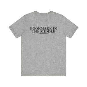 Puede incluir: Una camiseta de manga corta en color gris jaspeado con el texto negro BOOKMARK IN THE MIDDLE impreso en una fuente serif en el pecho, con una fina línea horizontal situada directamente debajo de la palabra MIDDLE.