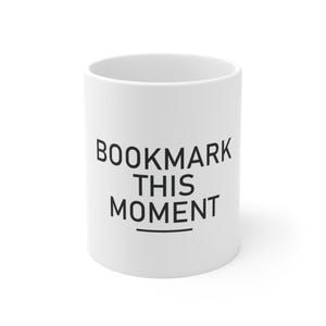 Peut inclure: Un mug en céramique blanche avec le texte noir BOOKMARK THIS MOMENT imprimé dans une police sans empattement sur le devant. Le mug présente une forme cylindrique avec une finition lisse et un design simple pour les boissons chaudes.