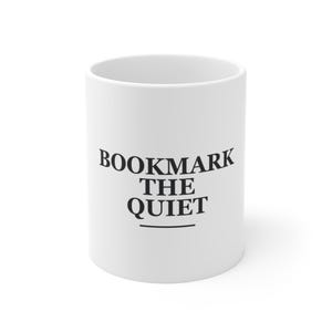 Peut inclure: Un mug en céramique blanche avec le texte noir BOOKMARK THE QUIET imprimé dans une police serif grasse sur le devant, accompagné d'une ligne horizontale sous le mot QUIET, centré sur la surface cylindrique lisse de l'objet.