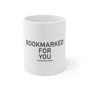 Peut inclure: Un mug en céramique blanche avec le texte noir BOOKMARKED FOR YOU imprimé dans une police sans empattement sur le devant. Le design comprend une ligne horizontale sous le mot YOU, centré sur la surface lisse et cylindrique.