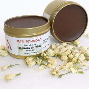 May include: A gold-colored tin of floral wax with the text "Jasmine Sambac" and "Altai Botanicals". The tin is open, revealing a dark brown wax. Dried jasmine flowers are scattered around the tin. The net weight is 2oz / 60g.