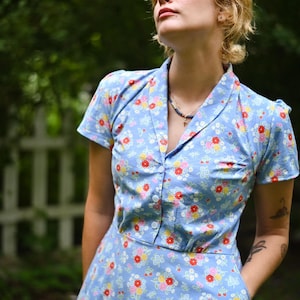 May include: A light blue short-sleeved dress featuring a repeating floral pattern in red, yellow, and white. The dress has a collared neckline, a button-down front closure, and visible side pockets. The fabric is a solid light blue cotton blend.