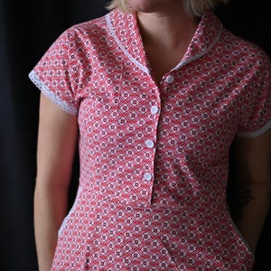 May include: A red and white patterned dress with short sleeves and a collared neckline. The dress features a geometric floral design and white lace trim on the sleeves and collar. It has white buttons down the front.