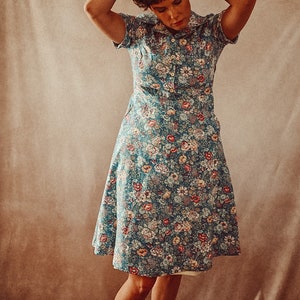 May include: A short-sleeved, button-down dress with a floral pattern in shades of blue, red, and cream. The dress has a collared neckline and a flared skirt. The person is wearing black shoes and gray socks. The dress is knee-length.