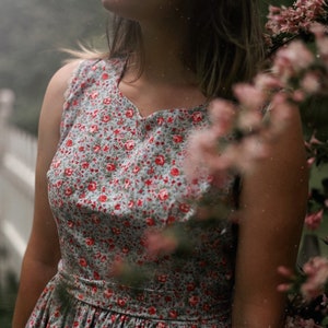 May include: A sleeveless dress with a light blue background and a red floral pattern. The dress has a fitted bodice and a flared skirt. The person is standing outside near pink flowers and a white fence.
