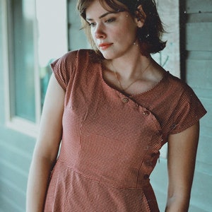 May include: A red and white checkered dress with short sleeves. The dress has a fitted bodice and a flared skirt. The neckline is asymmetrical with button details. The dress is made of a lightweight fabric, perfect for warm weather.
