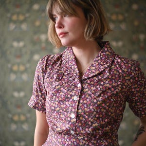 May include: A floral print dress with short sleeves and a collared neckline. The dress features a button-down front and a pattern of small flowers in shades of pink, yellow, and white on a dark background. The dress is worn by a person.