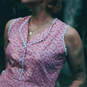 May include: A sleeveless pink dress featuring a small red and white floral pattern, a notched collar, and white lace trim along the edges. The bodice includes three light blue buttons down the front and a side zipper closure for a secure fit.
