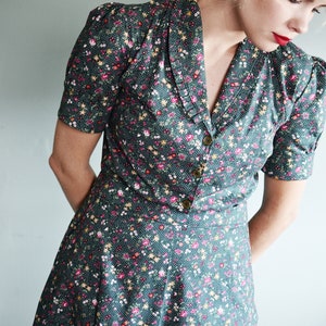 May include: A dark teal dress with a floral pattern featuring pink, yellow, and white flowers. The dress has short sleeves, a V-neckline, and a button-down front. The dress is a vintage style.