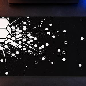 Puede incluir: Una alfombrilla de escritorio rectangular con fondo negro y un estampado gráfico blanco. El diseño consiste en un grupo de hexágonos en el lado izquierdo, que se transforman en formas hexagonales blancas dispersas y salpicaduras.
