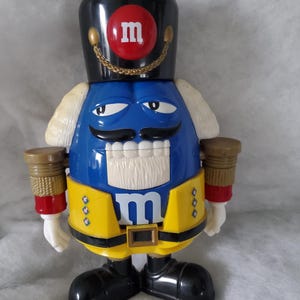 May include: A blue M&M nutcracker figure wearing a black hat with a red M logo, a yellow jacket with black trim, and black shoes. The figure features a white mustache, white hair, and a gold chain detail on the hat, standing in a rigid pose.