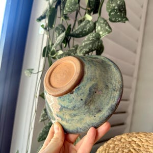 May include: A ceramic bowl featuring a mottled glaze in shades of blue, green, and hints of purple. The exterior surface displays a textured, variegated finish, while the circular base is left in an unglazed, natural terracotta clay color.