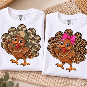 Puede incluir: Dos camisetas blancas con etiquetas Comfort Colors. La camiseta de la izquierda muestra un pavo marrón con una gorra de béisbol de camuflaje verde y plumas de cola de camuflaje. La de la derecha muestra un pavo marrón con un lazo rosa de lunares y plumas de cola con estampado de leopardo.