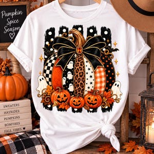May include: A white short-sleeve t-shirt featuring a large graphic of a pumpkin decorated with a black bow, leopard print, and checkered patterns. The design includes smaller jack-o'-lanterns, white ghosts, and autumn leaves on a black striped background.