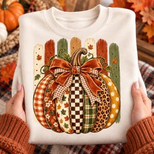 Op de afbeelding: Een crèmekleurig sweatshirt met ronde hals en een grote centrale afbeelding van een pompoen. De pompoen is opgebouwd uit verticale segmenten met diverse patronen, waaronder ruiten, bloemen, een dambordpatroon en luipaardprint, afgewerkt met een grote strik.