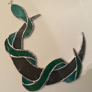 May include: A stained glass snake ornament featuring a curved body made of alternating dark green, teal, and translucent grey glass segments held together by silver-coloured metal solder lines, with a small metal loop at the top of the head.