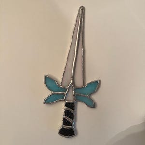 May include: A stained glass dagger ornament featuring a long, pointed white blade, a crossguard with two symmetrical light blue wings, and a black textured handle. The entire piece is outlined in silver metal solder, creating a defined shape.