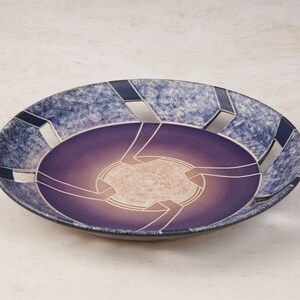 May include: A purple and blue ceramic bowl with a geometric design and cut-out details. The bowl has a speckled glaze and a white interior.
