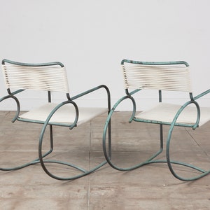Op de afbeelding: Twee turquoise metalen schommelstoelen met witte gevlochten zittingen en rugleuningen. De stoelen staan op een lichtgrijze betonnen vloer.