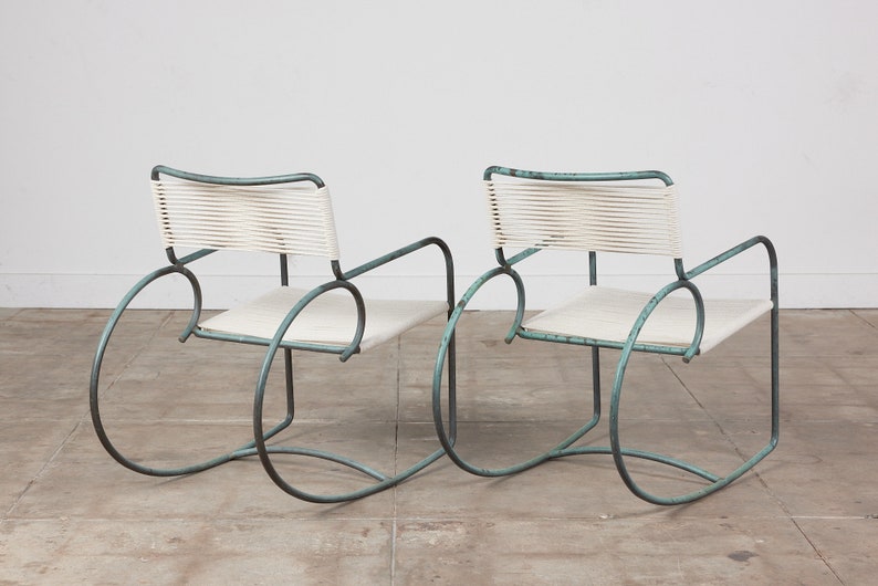 Op de afbeelding: Twee turquoise metalen schommelstoelen met witte gevlochten zittingen en rugleuningen. De stoelen staan op een lichtgrijze betonnen vloer.