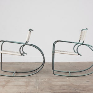 Op de afbeelding: Twee turquoise metalen schommelstoelen met witte gevlochten zittingen. De stoelen staan in een witte kamer met een betonnen vloer.