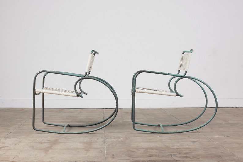 Op de afbeelding: Twee turquoise metalen schommelstoelen met witte gevlochten zittingen. De stoelen staan in een witte kamer met een betonnen vloer.