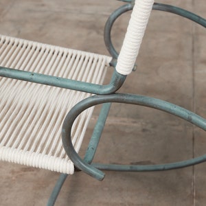 Op de afbeelding: Een close-up van een schommelstoel met een witte touwzitting en een groen metalen frame. De stoel is gemaakt van metaal en heeft een eenvoudig ontwerp.
