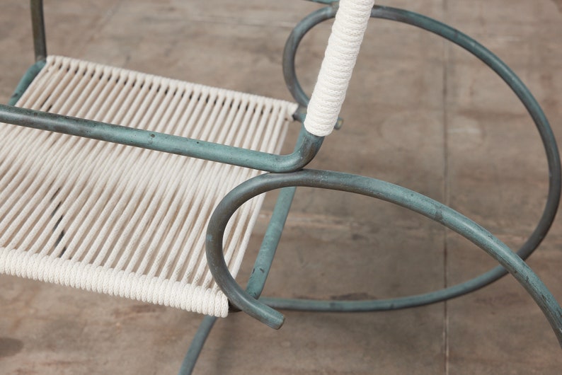 Op de afbeelding: Een close-up van een schommelstoel met een witte touwzitting en een groen metalen frame. De stoel is gemaakt van metaal en heeft een eenvoudig ontwerp.