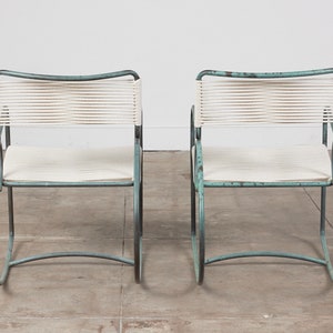 Op de afbeelding: Twee turquoise metalen stoelen met witte touwzittingen. De stoelen hebben een eenvoudig, modern ontwerp en zijn perfect voor buiten dineren of loungen.