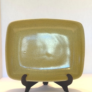 May include: A rectangular ceramic plate with rounded corners and a speckled chartreuse glaze. The plate is displayed upright on a dark brown, three-legged plastic stand, showcasing its smooth, glossy surface and slightly concave center.